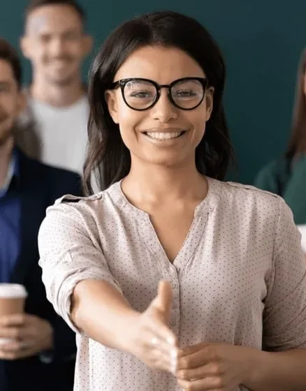 Smiling woman with glasses extending her hand for a handshake, with a group of colleagues in the background, suggesting a welcoming and professional environment.