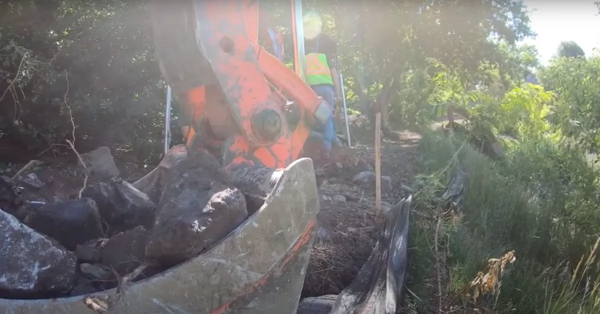 Excavator bucket filled with large rocks, working on a rocky terrain with workers in safety gear nearby in a wooded area.