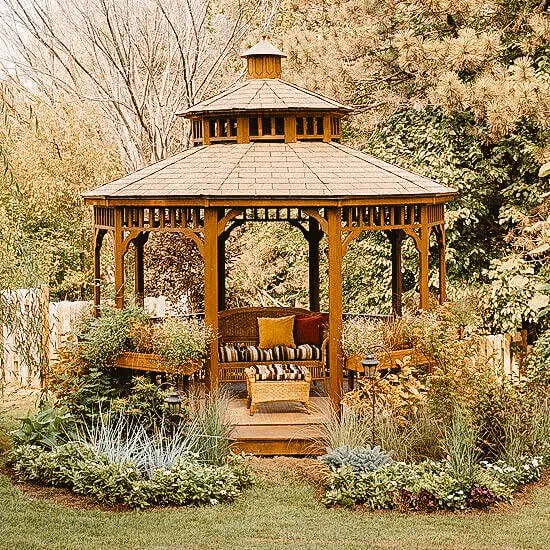 Wooden gazebo with a shingled roof, surrounded by lush greenery and garden plants, featuring comfortable seating inside.