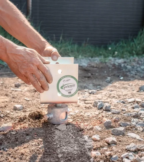 Hands adjusting a thermal pile foundation bracket on rocky ground, showing a close-up of the installation process.