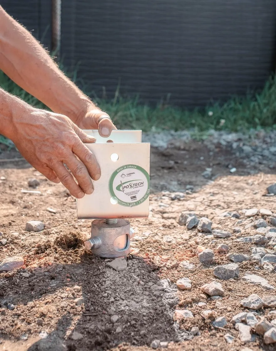 Hands adjusting a thermal pile foundation bracket on rocky ground, showing a close-up of the installation process.