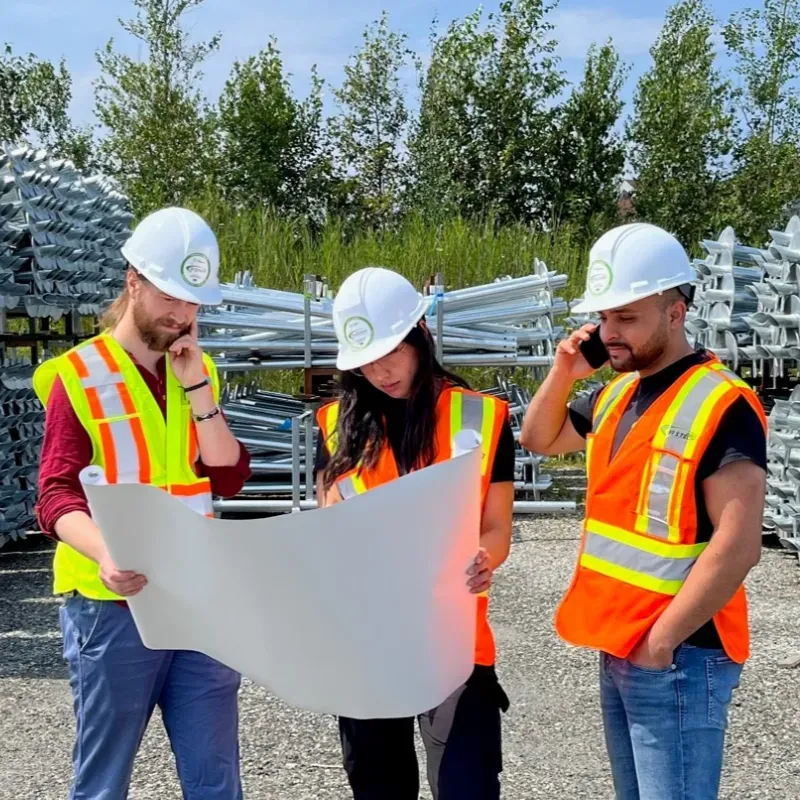 Three construction professionals wearing safety vests and hard hats, reviewing plans on a construction site, with piles of screw piles in the background.