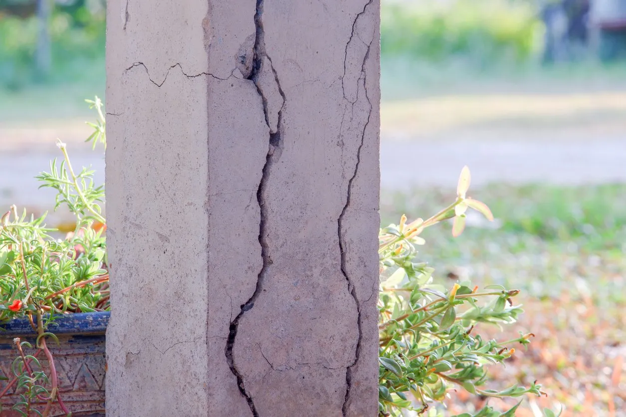 Cracked concrete foundation column with visible damage, surrounded by plants in an outdoor setting.