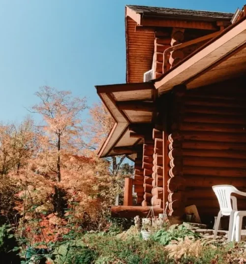 Log cabin nestled in a mountain forest with autumn-colored trees, featuring a small patio with white chairs under a clear blue sky.