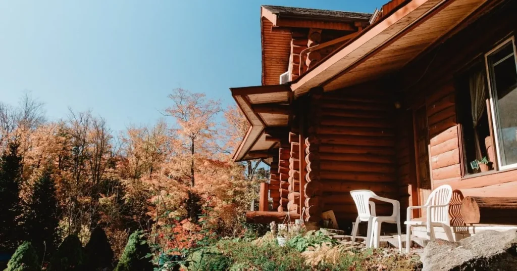 Log cabin nestled in a mountain forest with autumn-colored trees, featuring a small patio with white chairs under a clear blue sky.